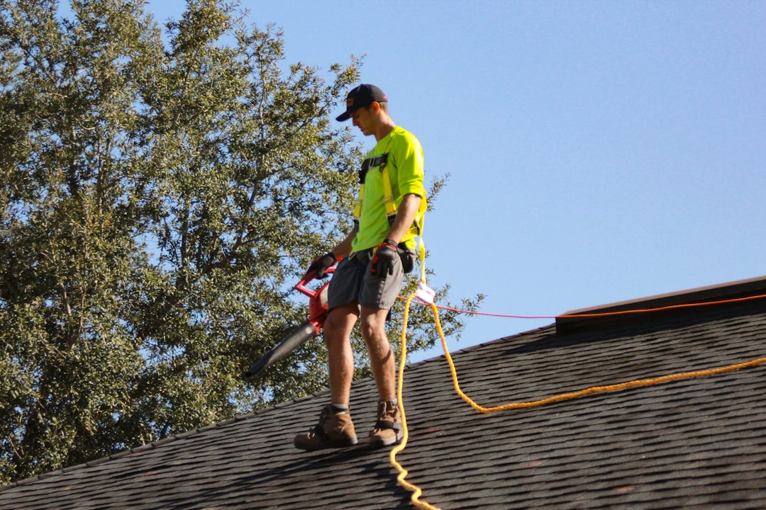 A man on a sloped shingled roof wearing a safety harness and holding a handheld tool