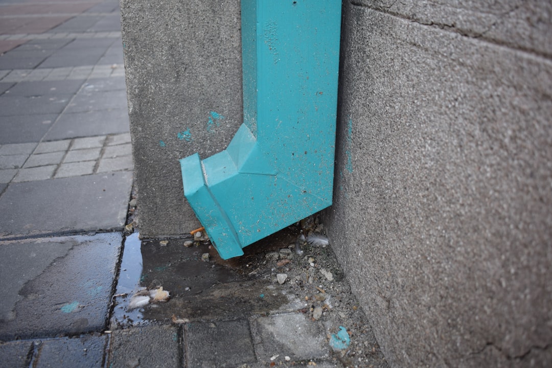 102d67bcb49745b7aa620ea601c1ea8a Teal downspout on a concrete wall, with wet ground below.