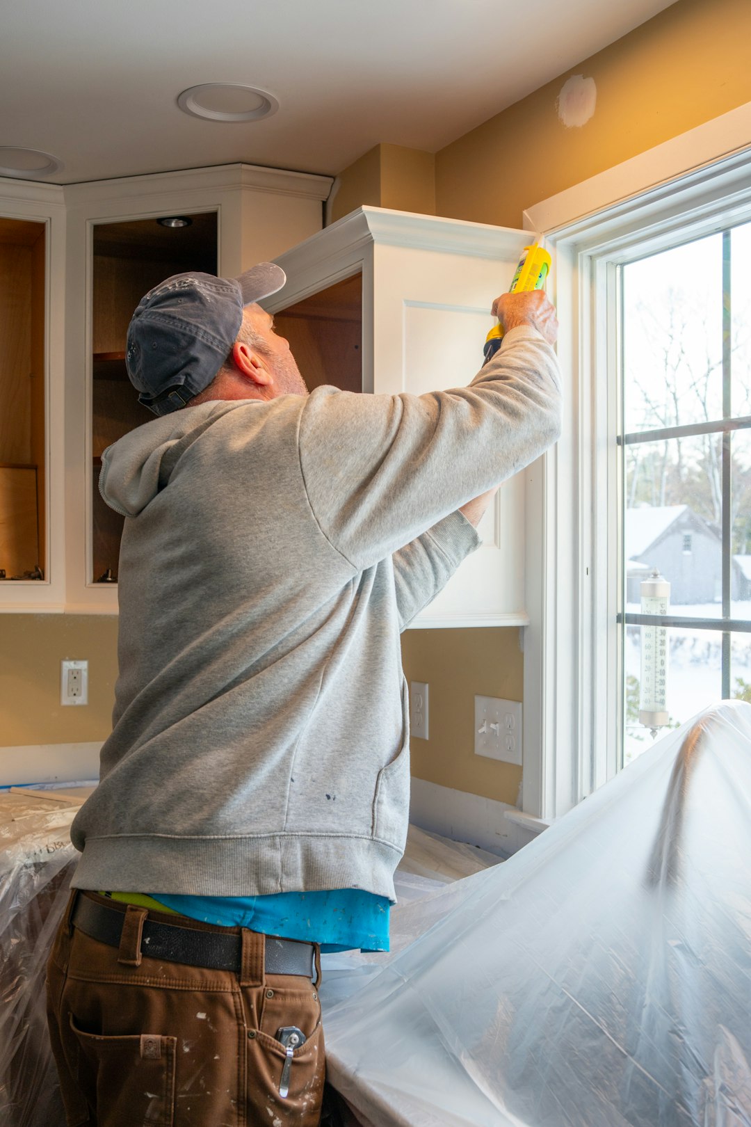 A person in a grey hoodie applying caulk to a window frame in a kitchen.