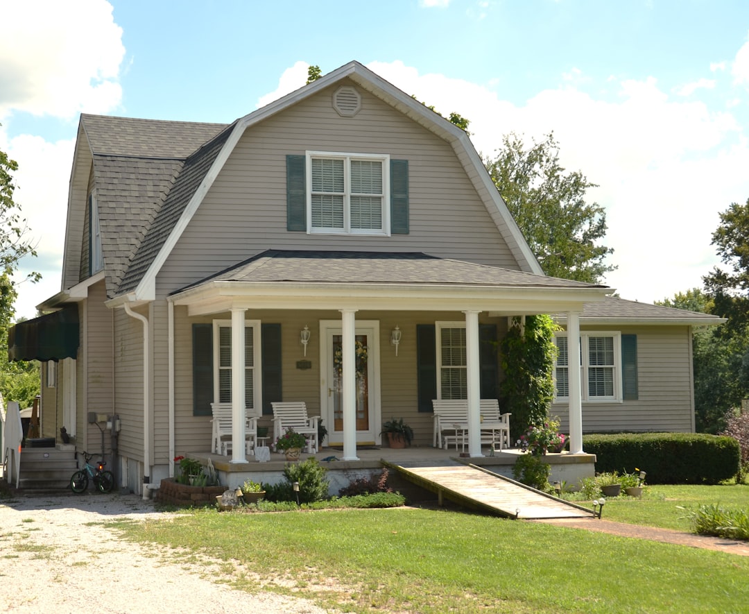 37d93b917d1547e98c50a63dbdf86f3f A beige house with a gambrel roof and a front porch with white columns
