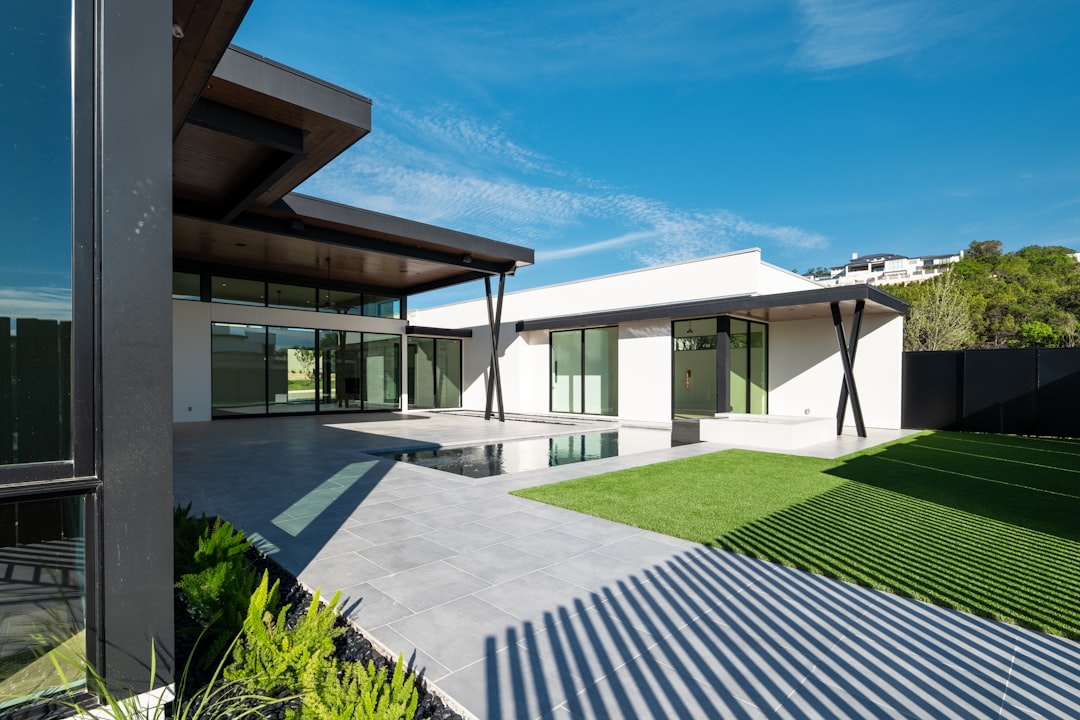 387b59c3dd1d4730b6a72f2964ca8338 Modern house with patio, green lawn, and blue sky.
