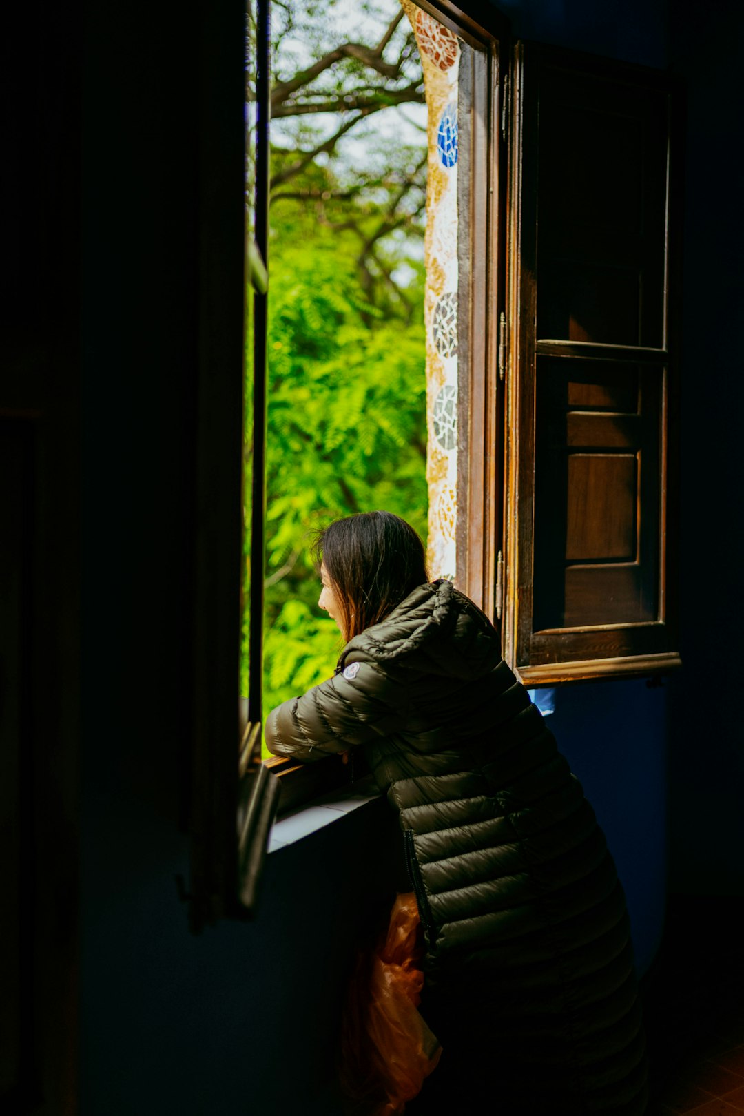A woman in a jacket looks out a bright window.