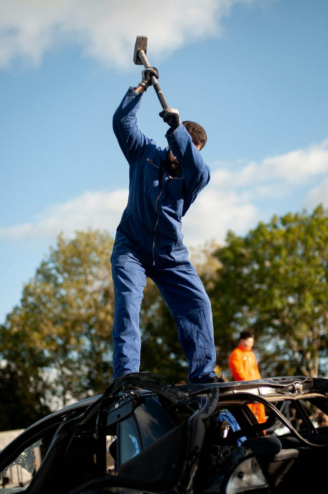 A man in a blue coverall raising a sledgehammer while standing on a wrecked car outdoors