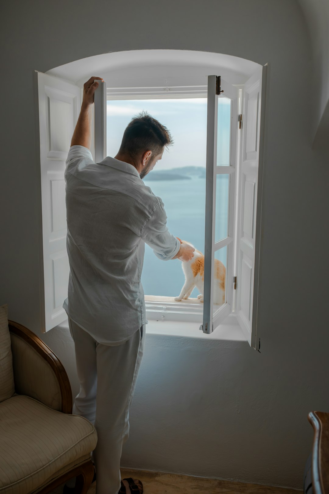 Man in white pets cat at window overlooking ocean.