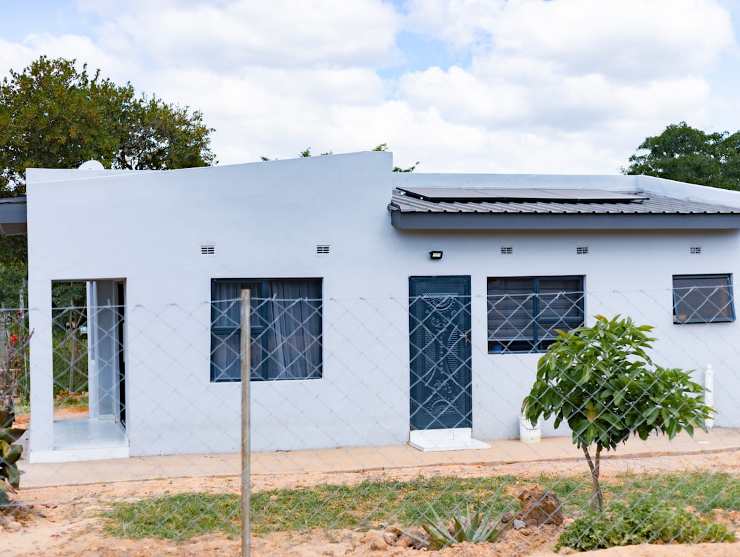 05449054a92945cba55c58d24977347b A small grey house with solar panels on the roof behind a wire fence