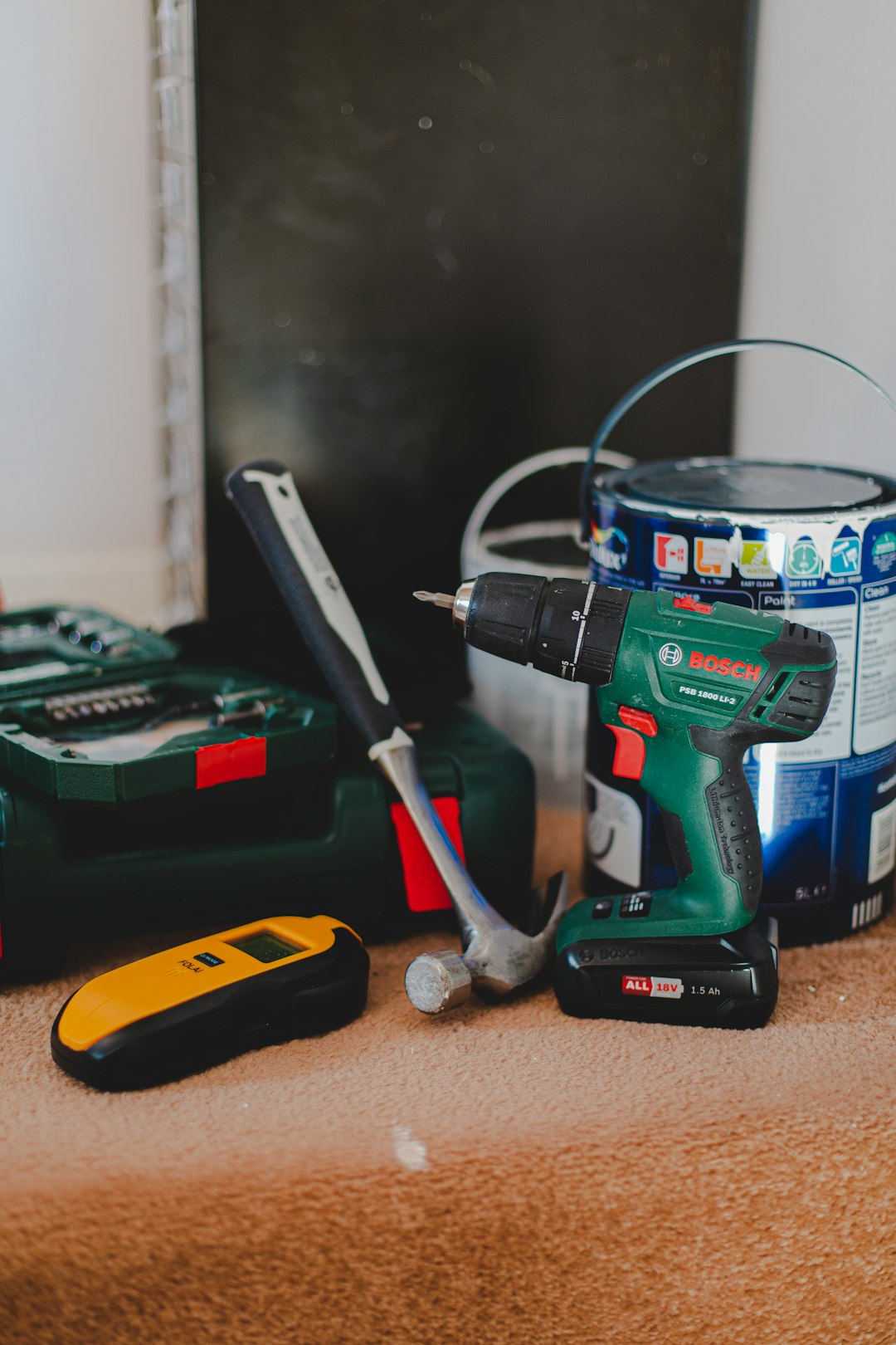 A cordless drill, a hammer, and a yellow stud finder on a carpeted floor